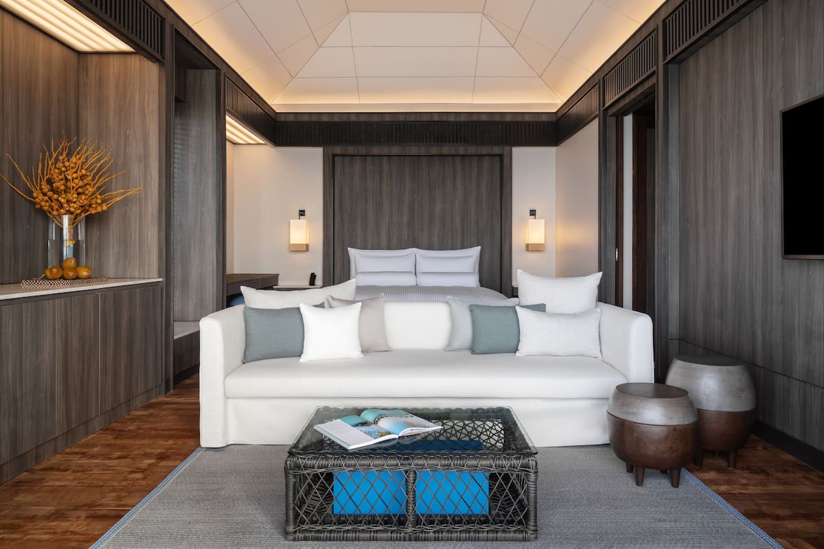 A modern bedroom with a white sofa, a large bed, a wicker coffee table, and two wooden side tables.