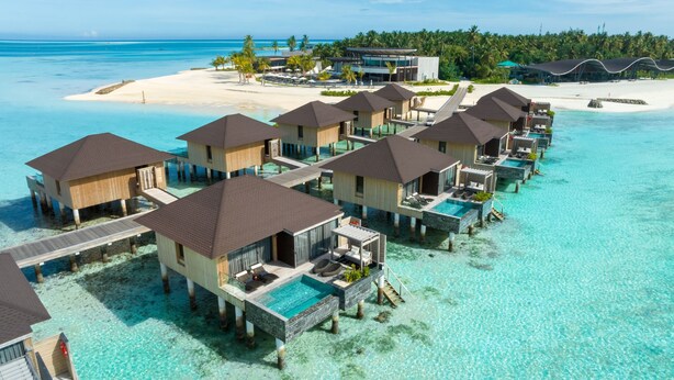 Overwater bungalows with private pools, connected by walkways, on a tropical island with clear turquoise waters and a sandy beach.