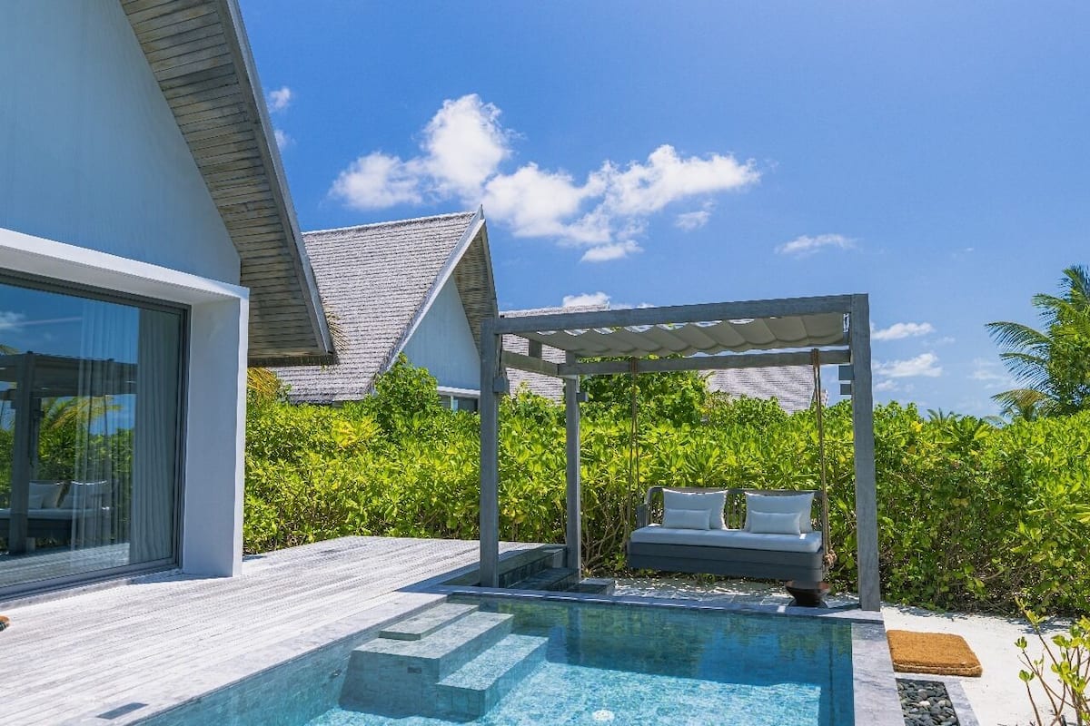 A modern pool area with a glass-enclosed room, a covered seating area, and lush greenery.