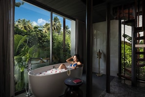 A woman relaxing in a bathtub with a glass of wine, large windows, and a view of tropical foliage.
