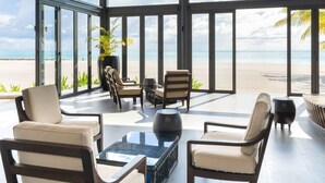 A modern lounge with large windows overlooking a beach, featuring comfortable seating and a glass coffee table.