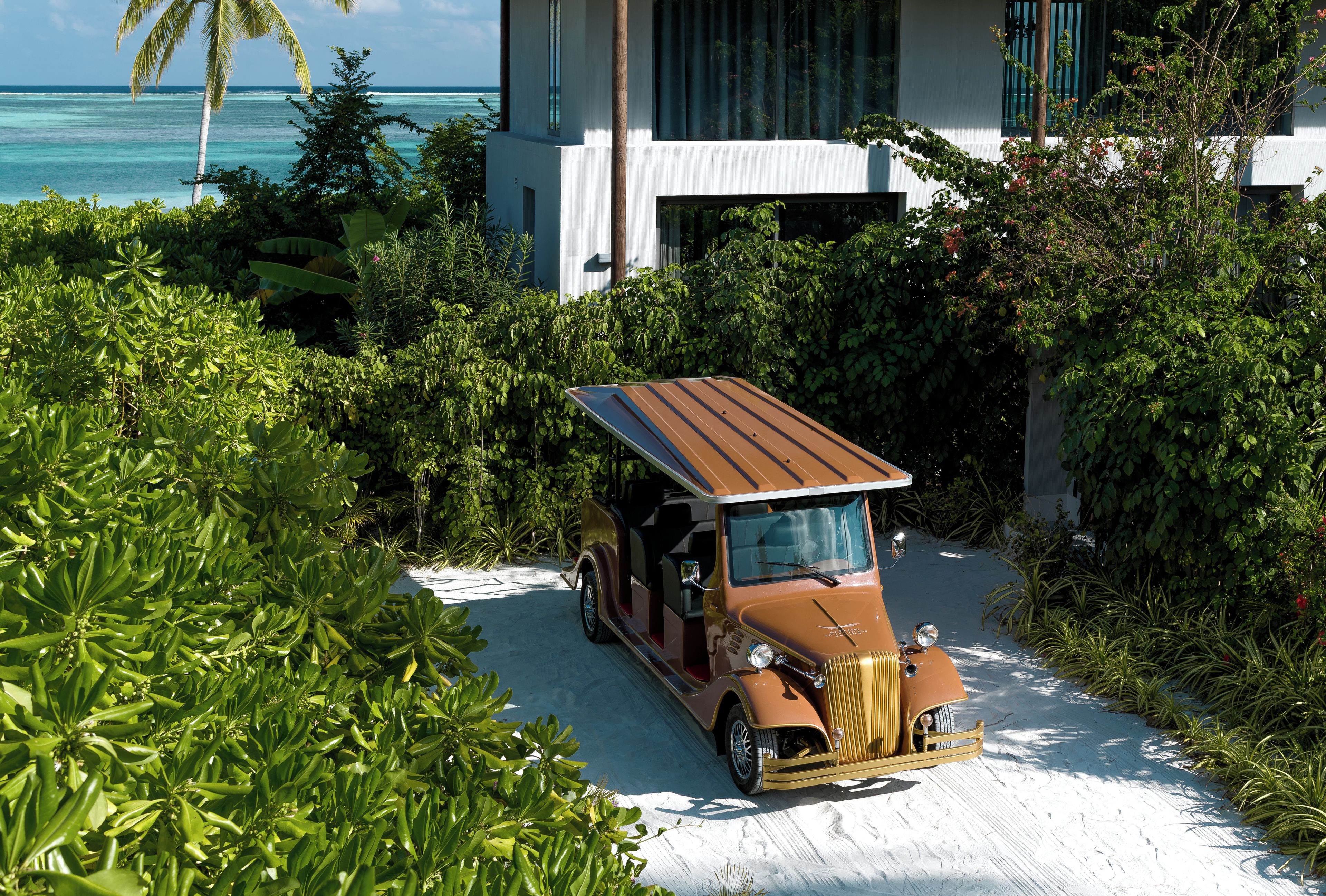 A vintage car parked in a lush garden with a modern house and ocean view in the background.