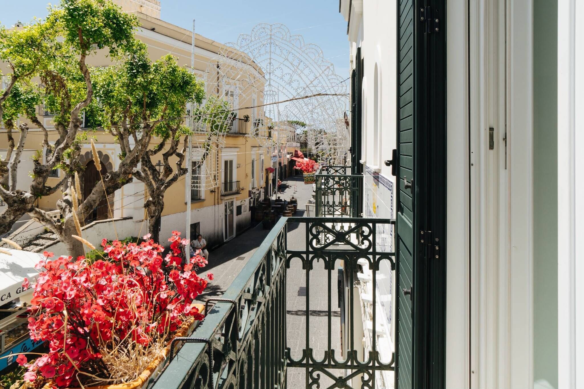 Een smalle straat met gebouwen aan weerszijden, een boom met rode bloemen op de voorgrond en een balkon met een metalen hekwerk.