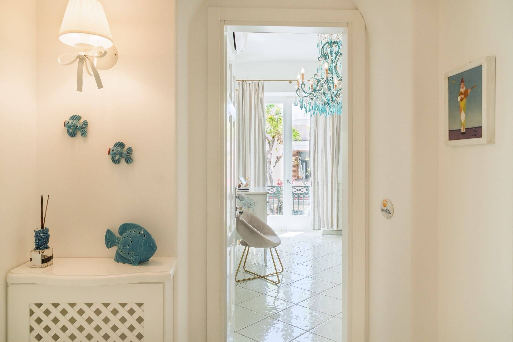 A hallway with a white console table, decorative fish, a wall-mounted lamp, and a door leading to a balcony with a chair and a hanging light fixture.