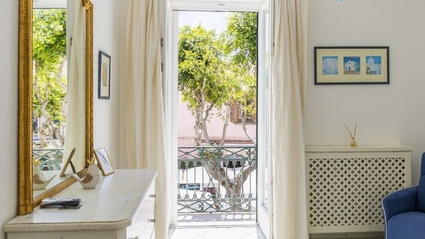 A room with a white dresser, a blue sofa, a chandelier, and a view of the outside. - Queen Suite (Forio)