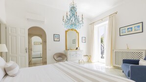 A hotel room with a large bed, a chandelier, a dresser, and a view of the outside. - Queen Suite (Forio)