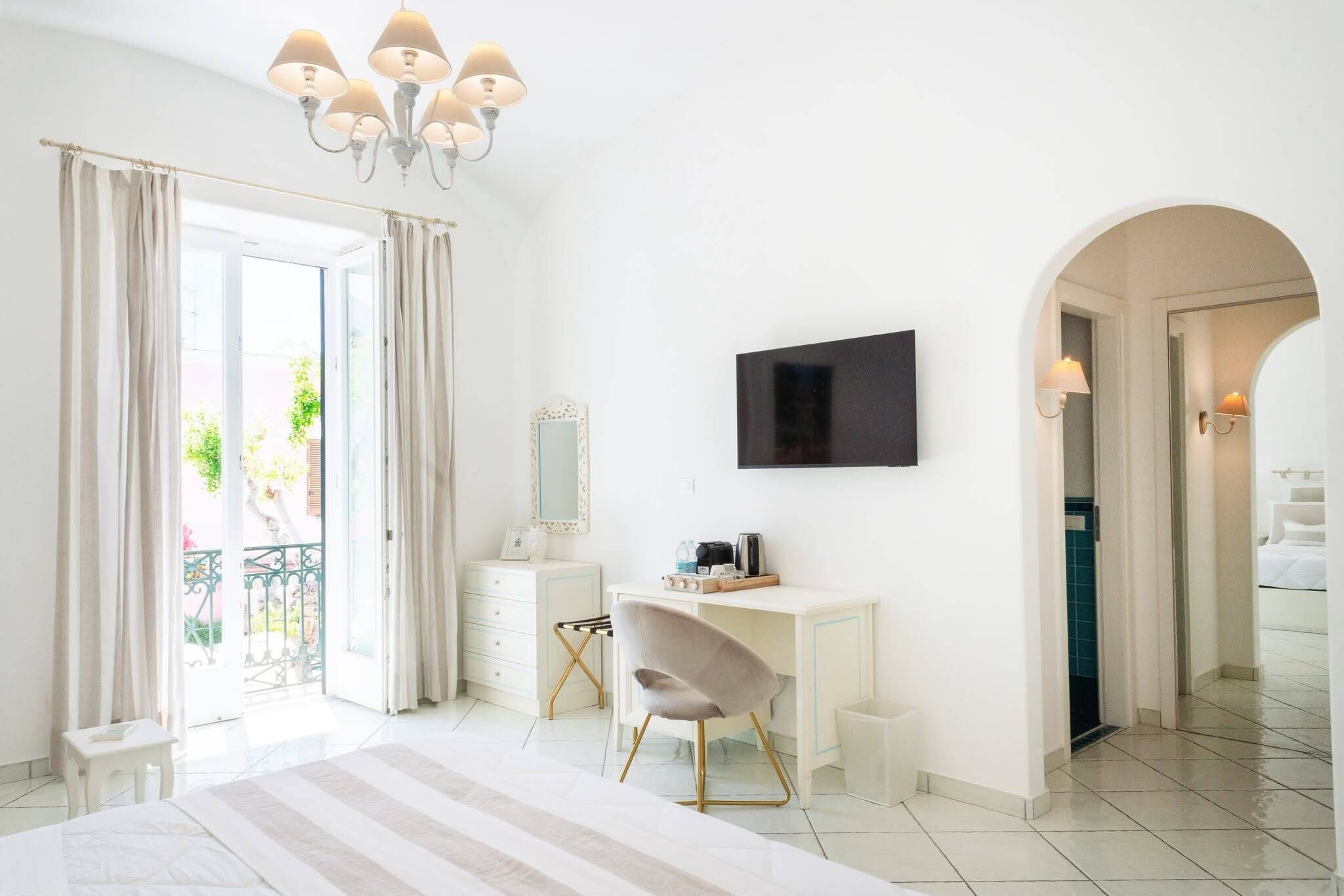 A modern bedroom with a large bed, a desk with a chair, a TV mounted on the wall, and a balcony visible through the door.