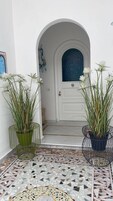 Uma porta branca com uma janela de vidro azul, acompanhada por dois vasos com plantas e flores brancas.