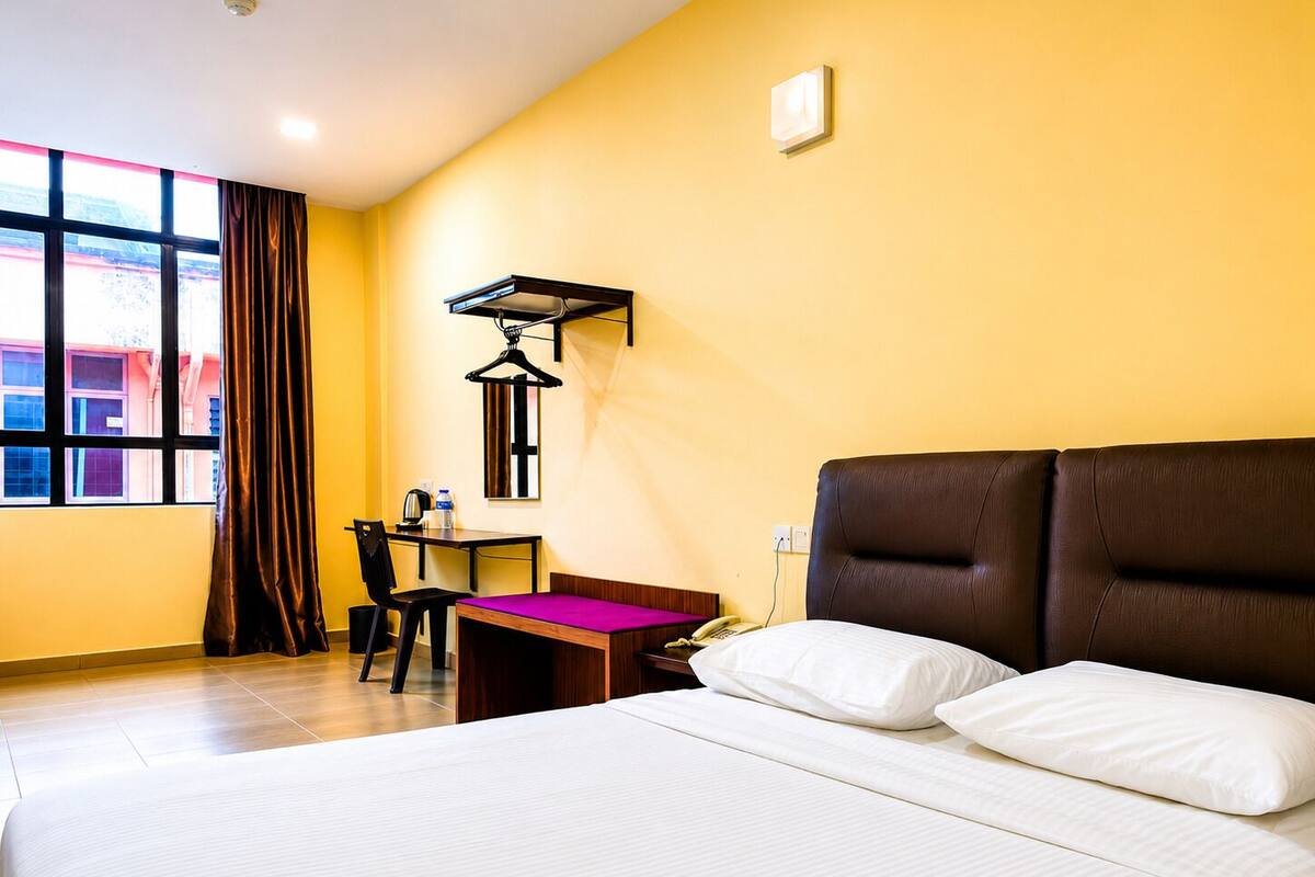 Habitación de hotel con cama, escritorio con silla, ventana con cortinas y lámpara fijada a la pared.