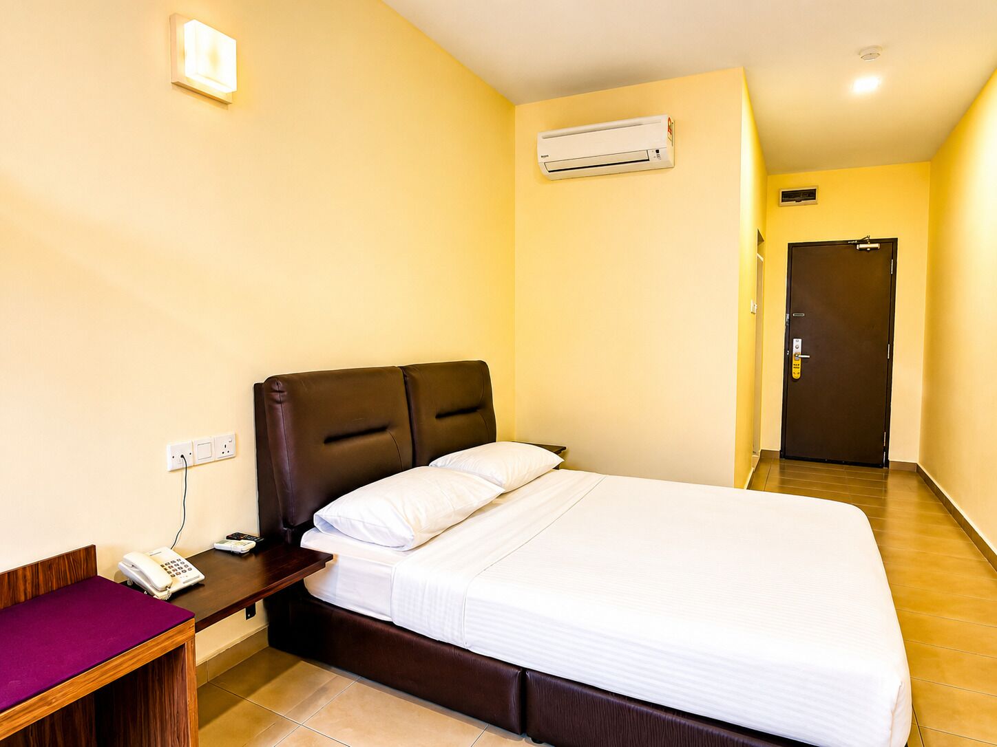 A hotel room with a bed, bedside table, phone, and an air conditioning unit on the wall.