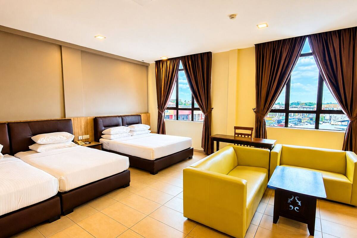Habitación de hotel con dos camas, un sofá, una mesita y vistas a la ciudad a través de amplios ventanales.