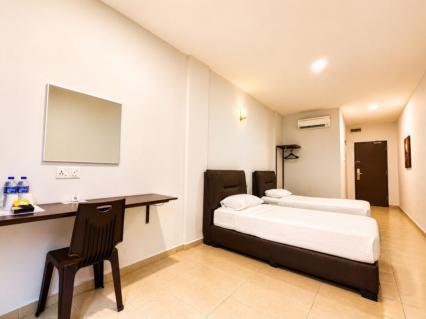 Habitación de hotel con escritorio, dos camas, una silla, un espejo, unidad de aire acondicionado y una puerta.