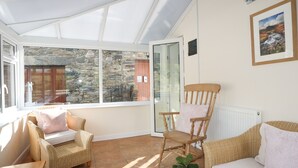 A sunlit room with wicker furniture, a wooden rocking chair, and a view through a glass door to an outdoor area with stone walls.