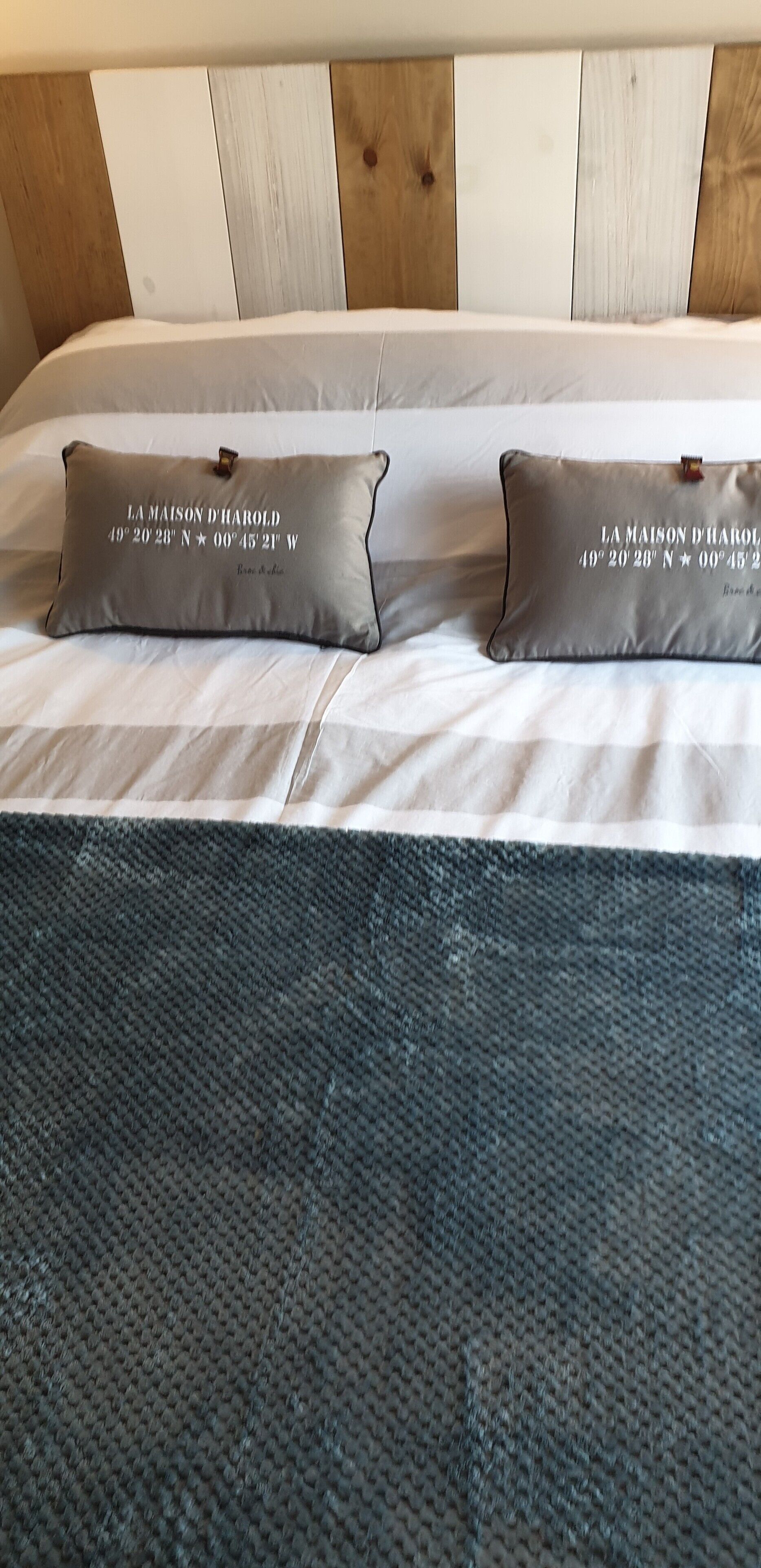 Een bed met twee kussens, een grijze geweven plaids en een houten hoofdbord.