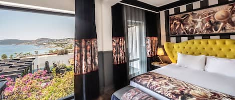 A hotel room with a large bed, a yellow tufted headboard, a view of the sea, and a balcony with flowers.
