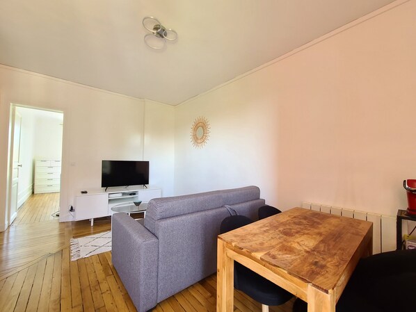 Una sala moderna con un sofá gris, una mesa de comedor de madera, un televisor de pantalla plana sobre un mueble blanco y un elemento decorativo en la pared.