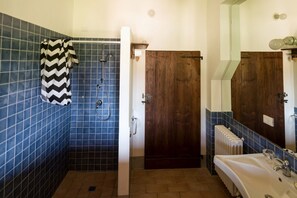 Ein Badezimmer mit blauen Fliesen, einer Dusche inklusive schwarz-weiß gestreiftem Handtuch, einer Holztür, einem weißen Waschbecken mit Spiegel und einem Heizkörper.