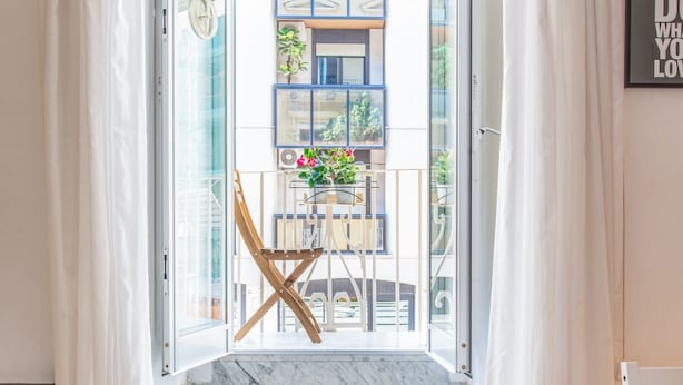 Une porte ouverte donnant sur un balcon où se trouvent une chaise, une table et des plantes en pot.