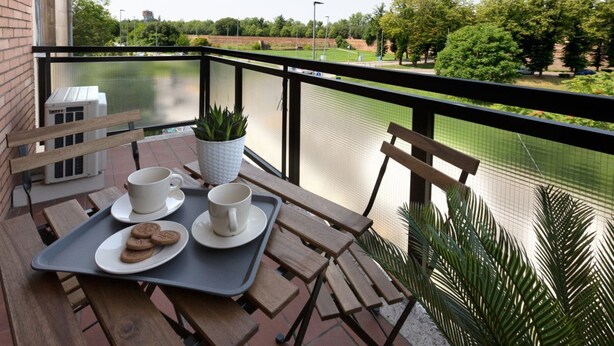A balcony with a table set for two, a potted plant, and a view of a body of water and greenery.