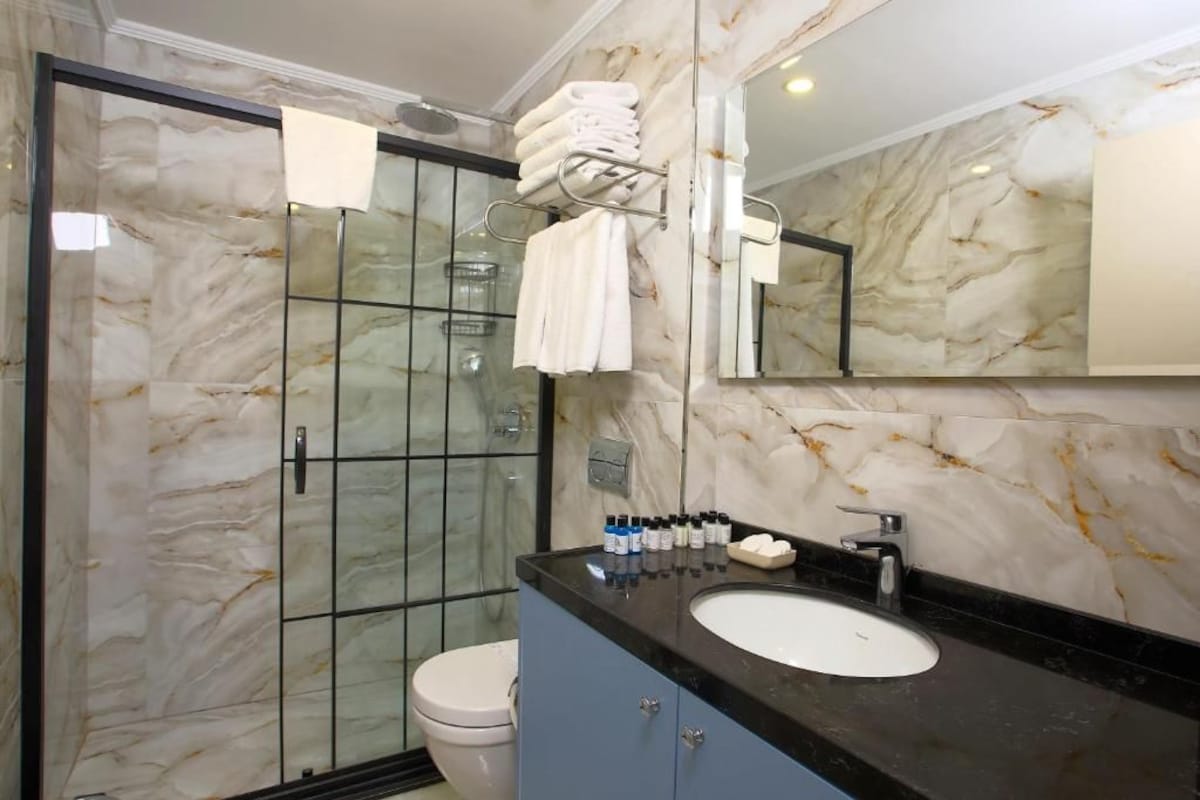 A bathroom with a marble wall, a glass shower enclosure, a black countertop, and a white sink.