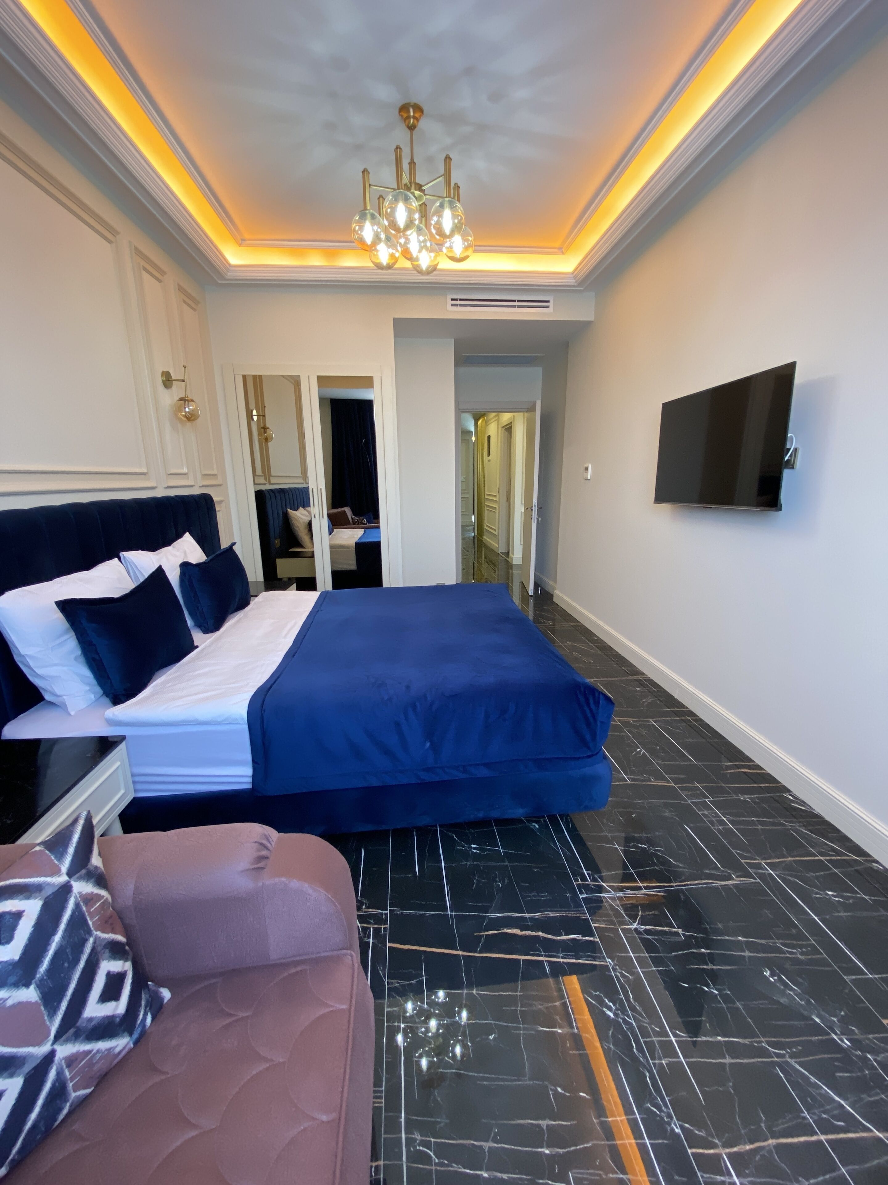 A modern hotel room with a large bed, a sofa, a TV mounted on the wall, and a chandelier.