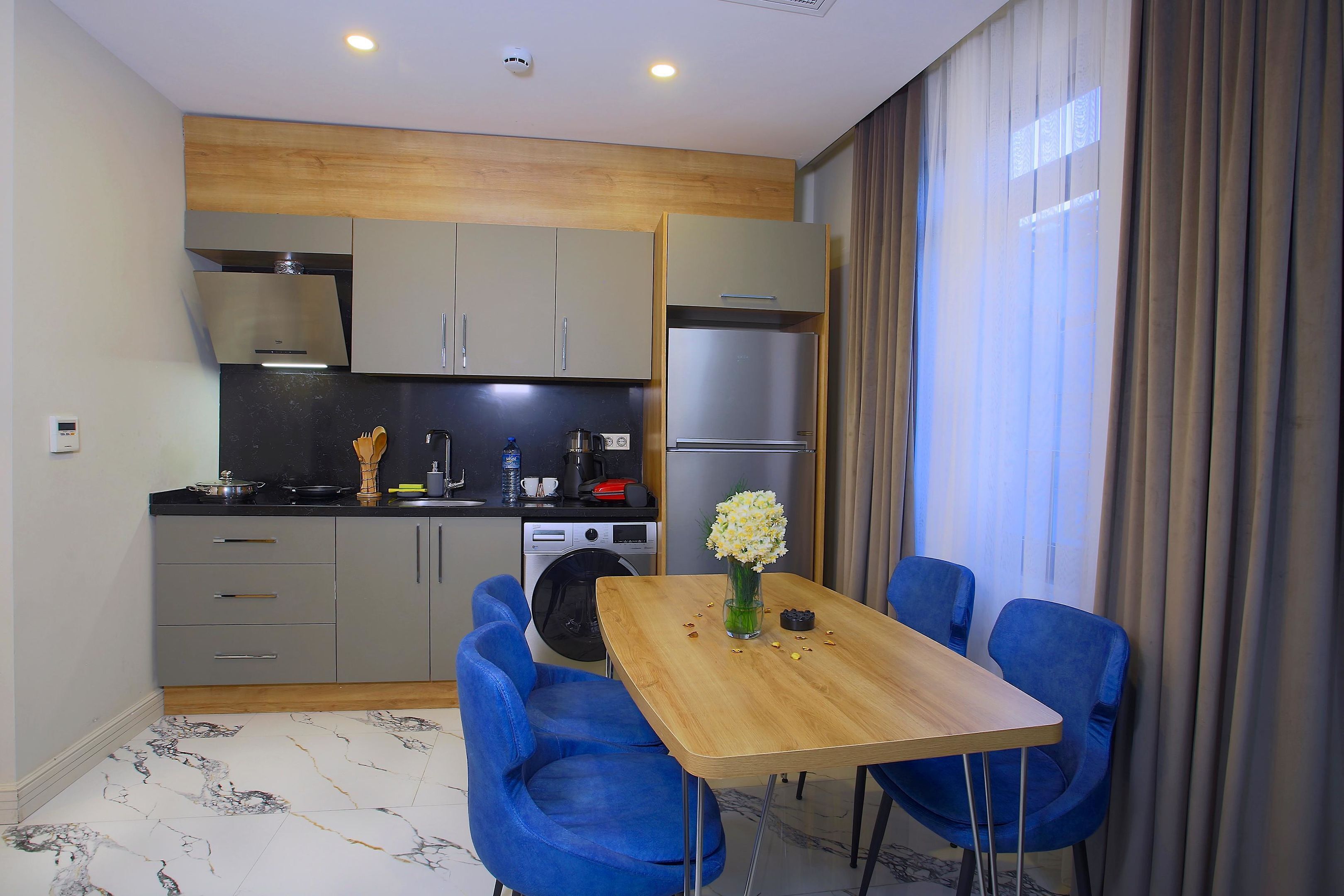 Una cocina compacta con electrodomésticos modernos, una zona de comedor con sillas azules y un amplio ventanal con cortinas.
