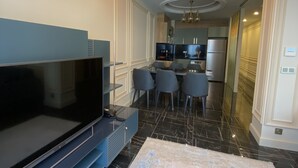 A modern living room with a large flat-screen TV, a dining area with chairs, and a kitchen in the background. - Princely Suites (Istanbul)