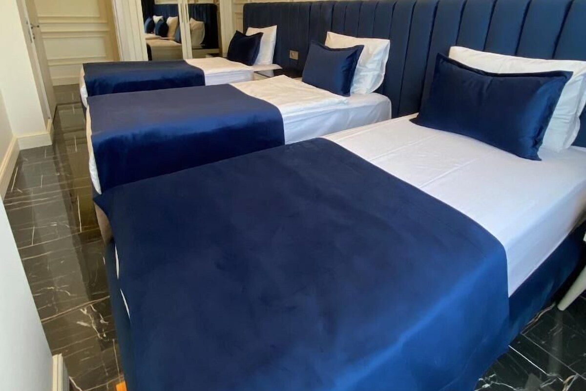 A hotel room with three beds, each with a blue bedspread and white linens. The room has a modern design with a marble floor and a chandelier.