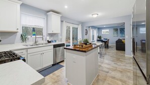 A modern kitchen with white cabinets, a central island, and a dining area with a table and chairs. - 5BR New Buffalo Home, Hot Tub, Fire Pit, Near Downtown (New Buffalo)