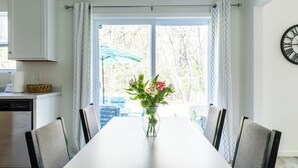 A dining area with a white table, chairs, a vase with flowers, and a view through a window with curtains. - 5BR New Buffalo Home, Hot Tub, Fire Pit, Near Downtown (New Buffalo)