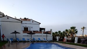 En swimmingpool med solstolar och parasoller, en terrass med kakelplattor och en byggnad med en vit fasad.