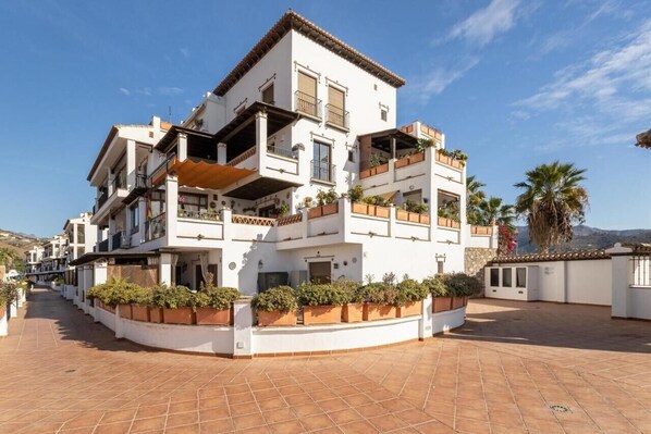 A multi-story residential building with balconies, a tiled courtyard, and surrounding vegetation.