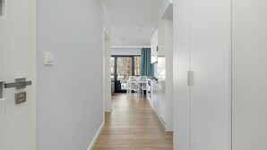 A narrow hallway with wooden flooring, white walls, and a dining area with white furniture in the distance.