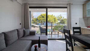 A modern living room with a sofa, dining table, and chairs, a kitchen area, and a large sliding glass door leading to an outdoor area with a hammock and greenery. - Villa with Sea View, Shared Garden & Wi-Fi (Lefkada)