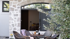 Un mur de pierres, des chaises en osier, une table avec des bouteilles de vin et une plante en pot.