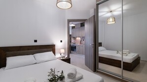 Une chambre d’hôtel moderne dotée d’un grand lit, d’une table de chevet, d’une lampe et d’un miroir avec une porte coulissante donnant sur une petite cuisine.