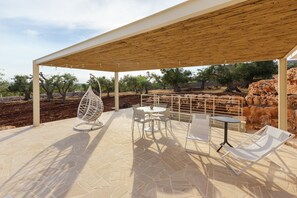 An outdoor patio with a wooden pergola, stone flooring, a hanging chair, a round table, and two lounge chairs.
