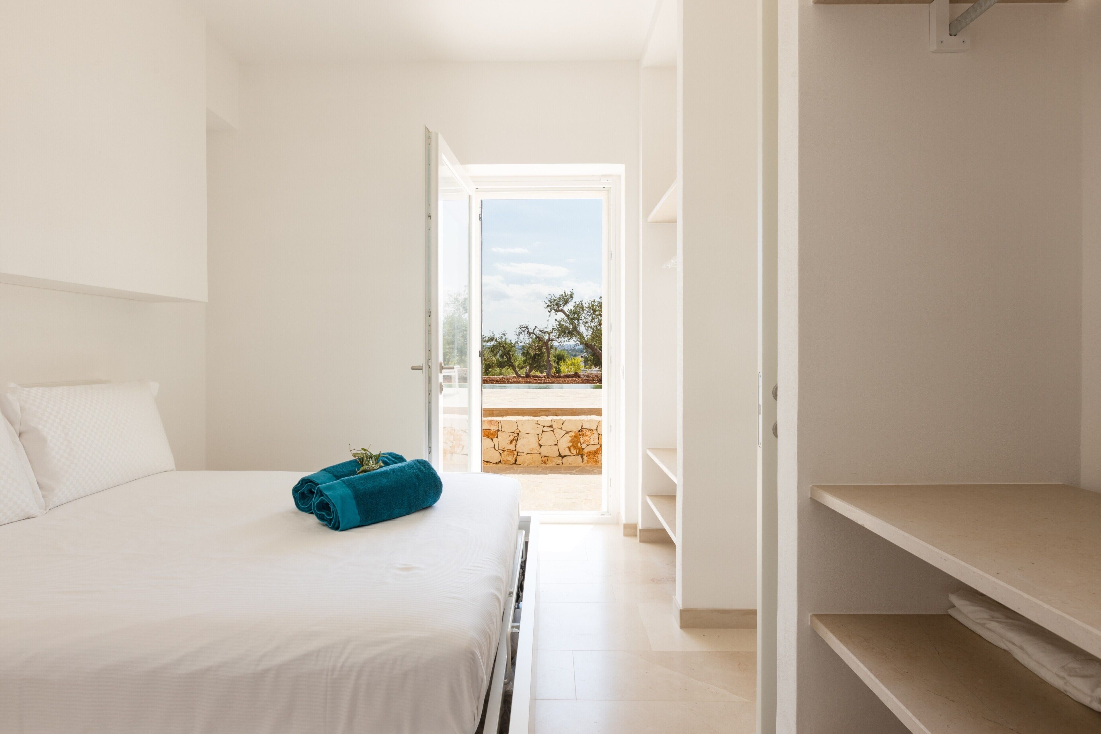 A hotel room with a bed, a blue towel, a white pillow, a wardrobe, and a view of the outdoors.