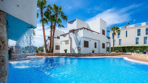 A swimming pool with a water feature, palm trees, and white buildings with balconies.