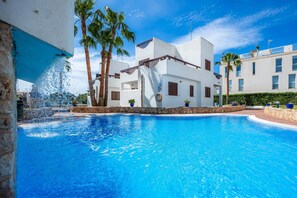 A swimming pool with a water feature, palm trees, and white buildings with balconies. - Apartment La Perla de Cala d'Or 3 with Shared Pool, Garden & Wi-Fi (Santanyí)