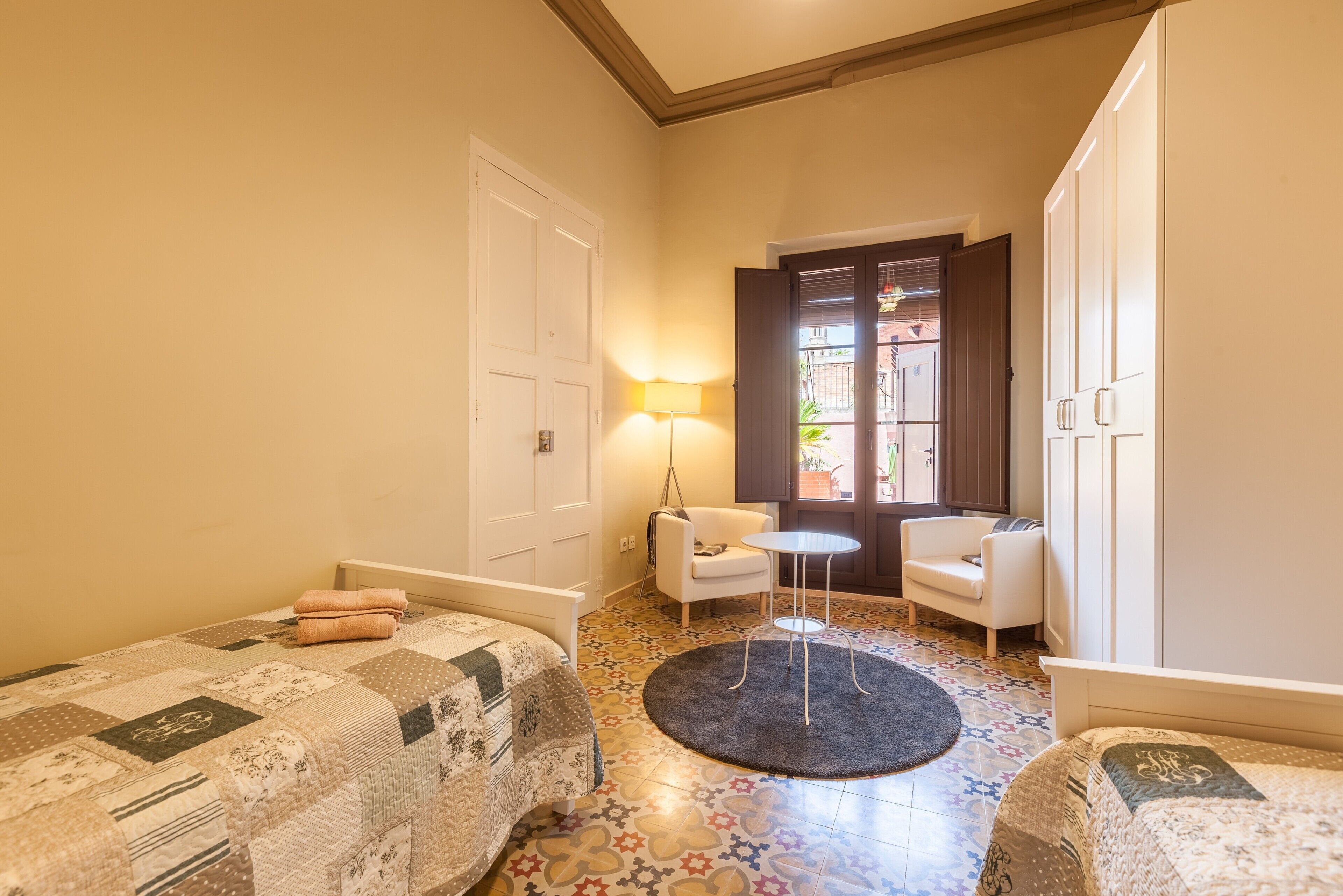 A hotel room with two beds, a small round table, a chair, and a door leading to a balcony.
