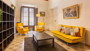 A living room with a yellow sofa, a matching armchair, a coffee table, a bookshelf, a window with blinds, and a painting on the wall.