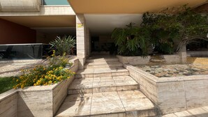 Une entrée d’immeuble avec un escalier recouvert de carreaux, un jardinier avec des fleurs jaunes et un grand arbre.