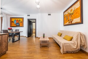 A room with a wooden floor, a sofa, a coffee table, a dining table with chairs, and two paintings on the walls.