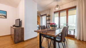 Un coin repas avec une table en bois, des chaises, un téléviseur posé sur un meuble et un balcon agrémenté de rideaux.