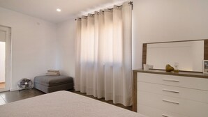 Un dormitorio con cama, mesita de noche, cómoda, espejo, ventana con cortinas y ventilador.