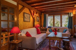 Interior de una cabaña de madera con sofá, mesa y ventana con vistas a árboles.
