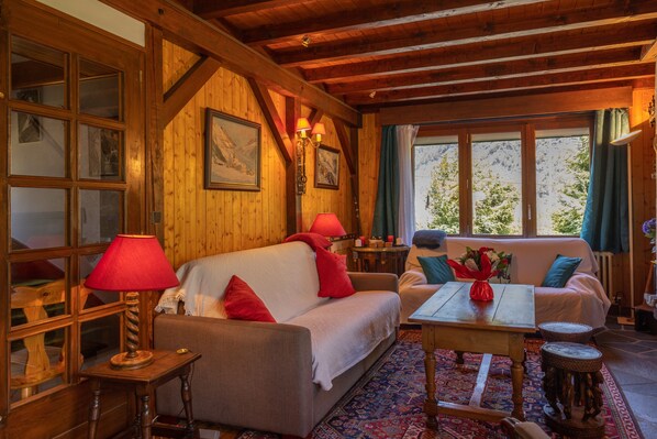 Interior de una cabaña de madera con sofá, mesa y ventana con vistas a árboles.