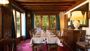 Une salle à manger avec une longue table dressée pour un repas, des chaises en bois, une moquette rouge et de grandes fenêtres avec des rideaux.