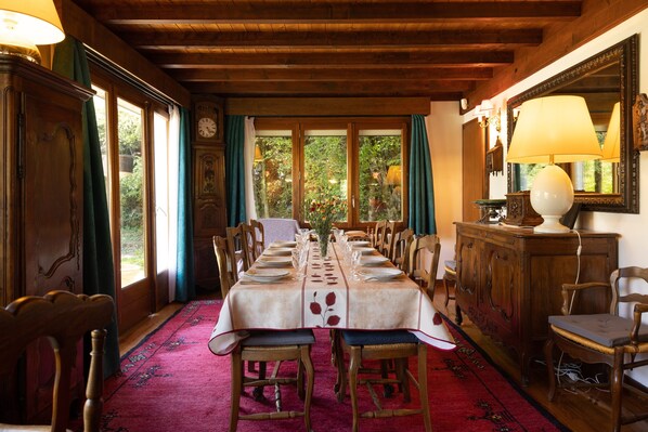 Une salle à manger avec une longue table dressée pour un repas, des chaises en bois, une moquette rouge et de grandes fenêtres avec des rideaux.